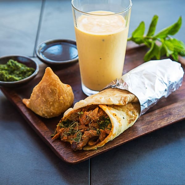 Kasa Indian Eatery's chicken kati roll, samosa, mango lassi, and chutneys on a wooden tray.