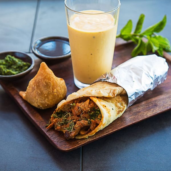 Kasa Indian Eatery's chicken tikka roll, samosa, mango lassi, and chutneys on a wooden tray.