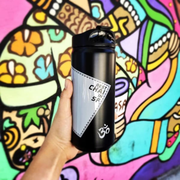 Hand holding a black Kasa Indian Eatery chai thermos against a vibrant, colorful mural.
