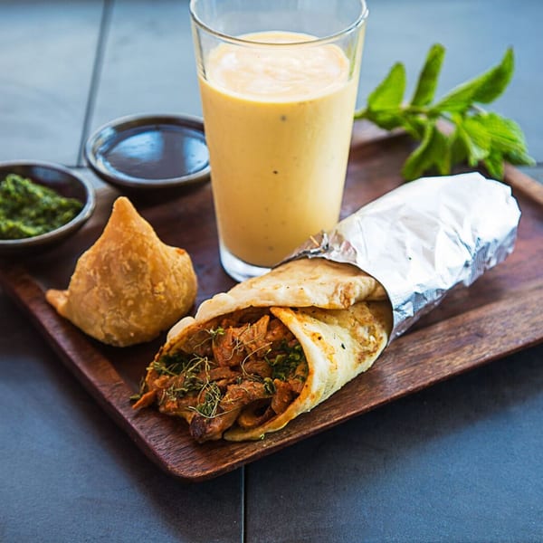 Kasa Indian Eatery's chicken tikka roll, samosa, mango lassi, and chutneys on a wooden tray.